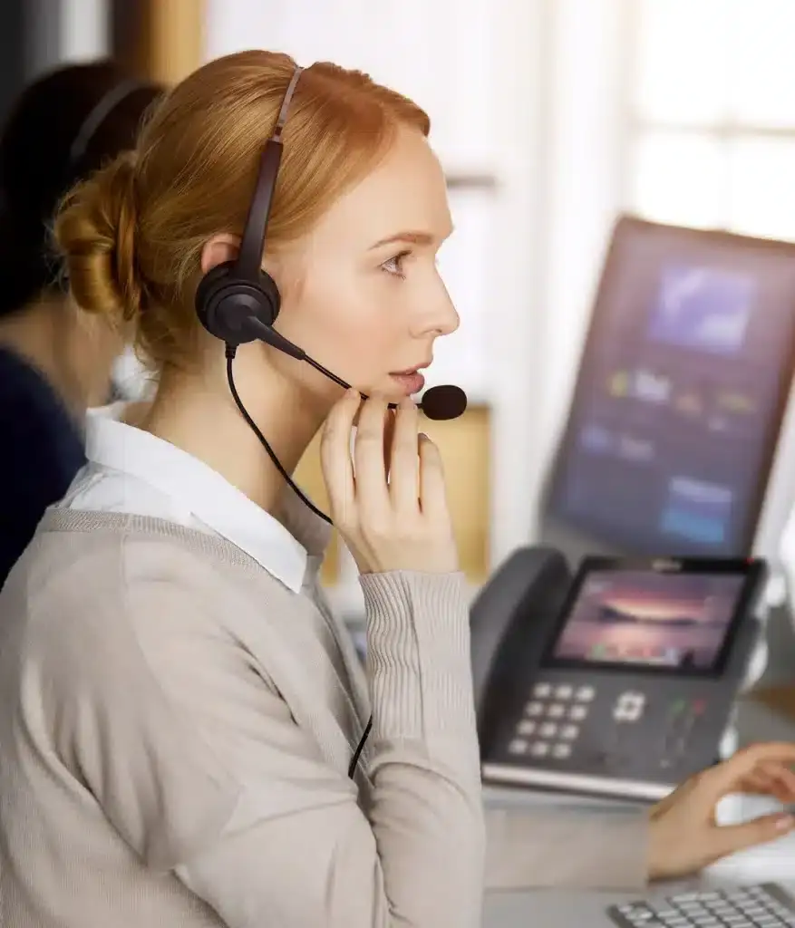 Call center agent using Ultatel cloud contact center solution, VoIP phone on the background and desktop computer showing the dashboard of the cloud contact center solution, used by another team member